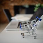 Carrinho de compras em miniatura sobre uma mesa ao lado de um laptop, representando compras públicas eletrônicas