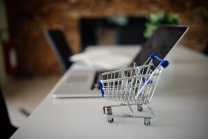 Carrinho de compras em miniatura sobre uma mesa ao lado de um laptop, representando compras públicas eletrônicas