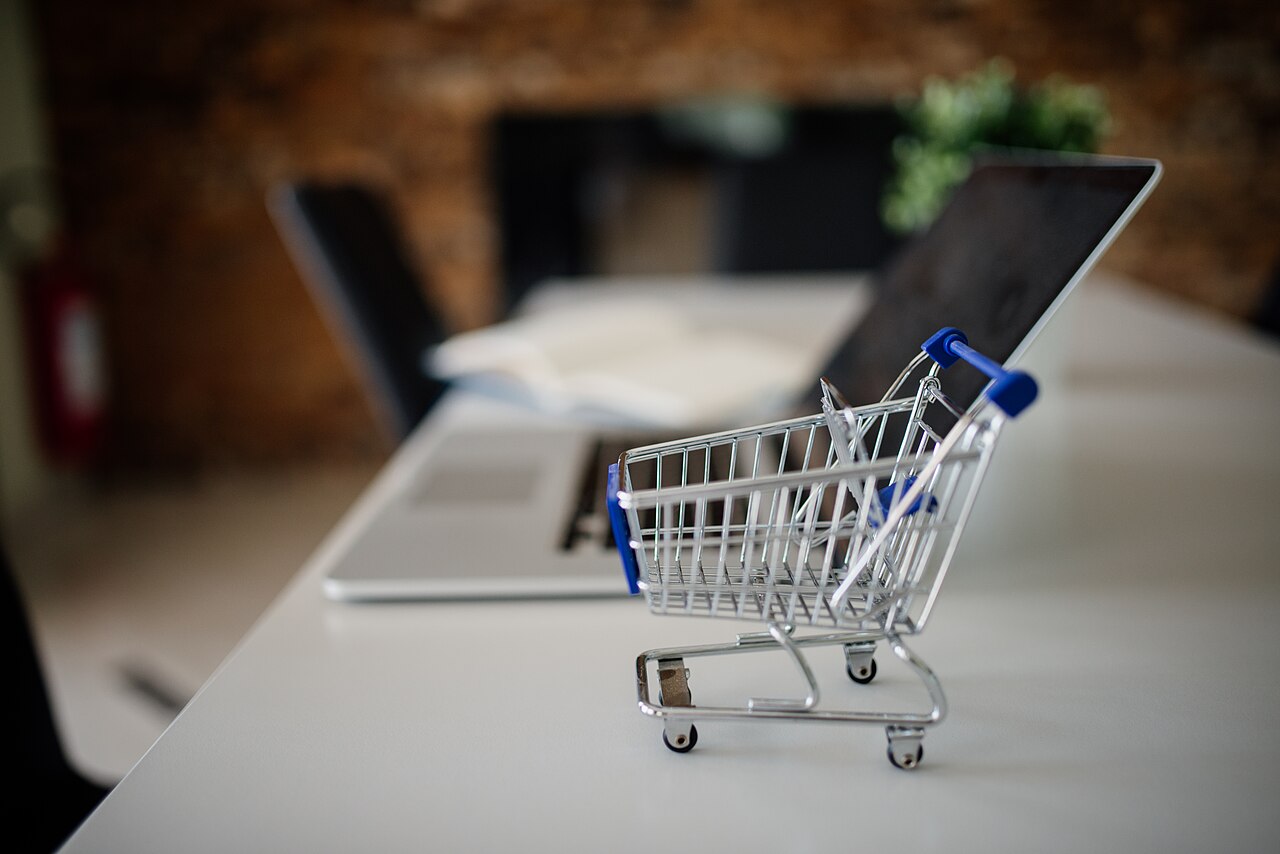 Carrinho de compras em miniatura sobre uma mesa ao lado de um laptop, representando compras públicas eletrônicas